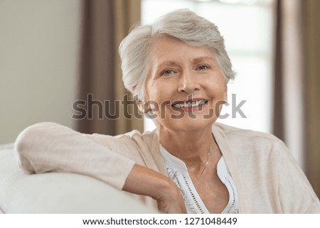 Smiling senior woman relaxing on couch at home and looking at camera. Portrait of elderly woman sitting on sofa. Closeup of cheerful grandmother relaxing indoor.
