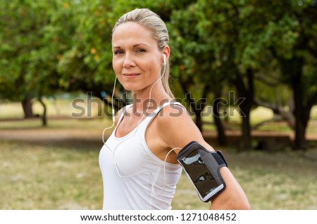 Portrait of athletic mature woman resting after jogging. Beautiful mid blonde woman running at park on a sunny day. Female runner listening to music while jogging and looking at camera.