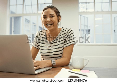 Laughing latin woman using laptop sitting on desk at office. Beautiful student finishing college assignments using computer in a library. Happy casual girl working on laptop and looking at camera.