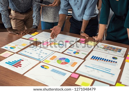 Financial and business documents on table with multiethnic hands working on it. Closeup business man and businesswoman hands understanding pie and bar graphs during meeting.