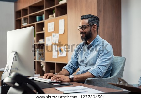 Smiling creative latin business man typing on desktop computer in office. Mature middle eastern businessman working at modern office space. Happy indian man wearing eyeglasses working on his computer.