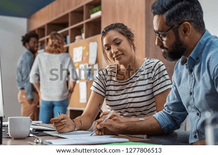 Mature businessman and young businesswoman discussing work and writing notes in office. Creative business partners working together sitting at desk. Middle eastern manager helping woman with details.