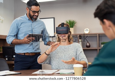 Mature executive man holding tablet showing video to woman wearing virtual glasses at office. Happy smiling client trying virtual reality in office with business team.
