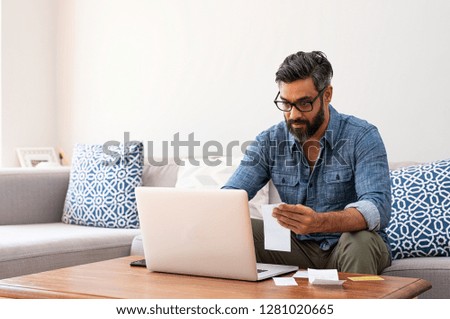 Mature man with spectacles and working on bills on laptop at home. Man with holding bills paying taxes with internet banking. Indian guy working on computer with invoice on table with copy space.