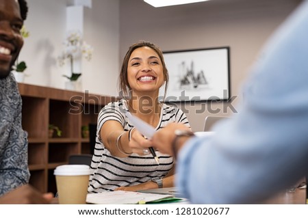 Smiling young businesswoman passing folder. Two multiethnic businesspeople sharing information sitting in meeting room with collegaues. Young beautiful businesswoman giving her work to executive.