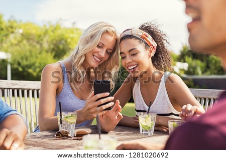 Two young women looking at smart phone and smiling sitting at table outdoor. Happy multiethnic girls taking selfie at outdoor cafeteria. Friends watching smartphone and laughing at park.