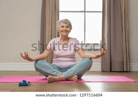 Senior smiling woman doing yoga in her living room. Elderly relaxed woman sitting in lotus pose and meditating while practicing yoga at home. Old grandmother sitting on yoga mat smiling.