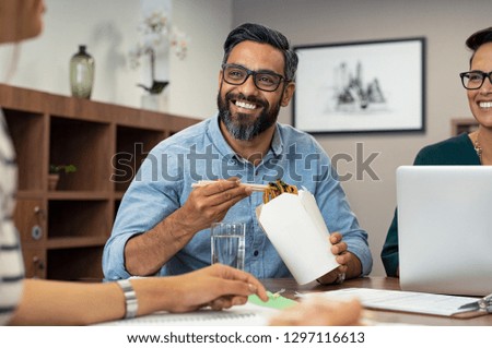 Multiethnic business man with colleagues having chinese take away food. Friendly businessman and casual businesswoman eating noodles. Mature latin man eating lunch meal while talking to creative team.