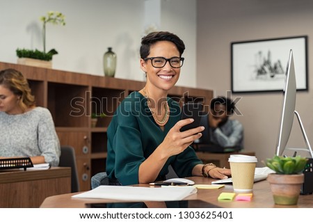 Happy fashionable girl using smartphone while working on desktop computer. Cheerful young business woman sitting in coworking space  and looking at camera. Portrait of  freelancer with eyeglasses.