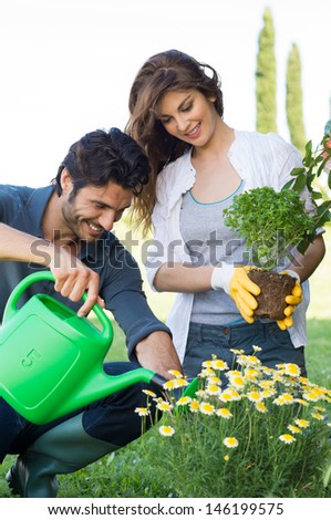 Woman Holding Plant Looking At Man Watering Plants In Garden