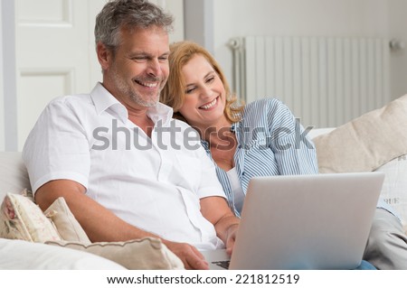 Portrait Of Happy Mature Couple Sitting On Couch Using Laptop