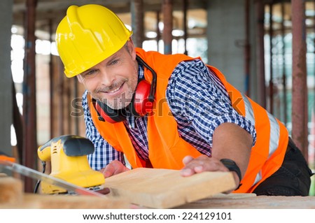 Close-up Of A Smiling Male Carpenter Looking At Wooden Plank At Construction Site
