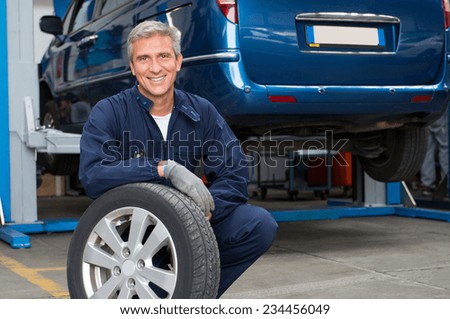 Portrait Of Happy Mature Mechanic At Repair Service Station Holding A Tyre