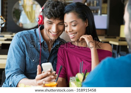 Closeup shot of young couple listening to music with mobile phone at coffee bar. Young man and african woman looking at smartphone at cafÃ?Â?Ã?Â©. Happy young couple listening music with headphones.