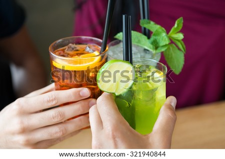 Closeup shot of friends toasting with cocktails. Young people drinking at aperitif. Shallow depth of field with focus on friends hand toasting juice glass. Close up of hands holding a cocktail glass.