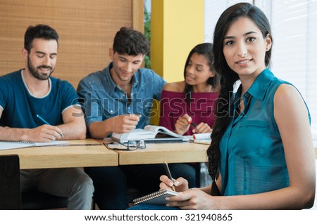 Portrait of young woman taking down note in notebook. Female student looking at camera and smiling. Happy student writing with other students studying in background.