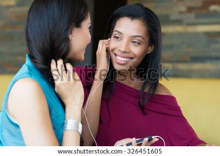 Closeup shot of young women listening to music with mobile phone. Happy female friends looking at eachother. Smiling girls listening to music indoor and smiling.