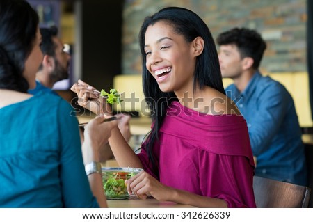 Closeup shot of young woman eating salad with her friend. African girl smiling at lunch. Lughing woman eating salada at restaurant during her lunch break.