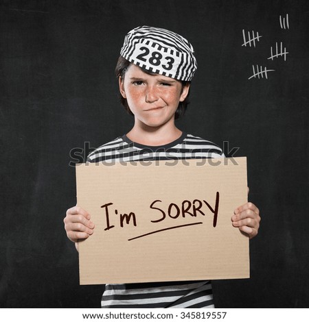 Closeup mugshot photo of boy holding I\'m sorry sign. Young boy make a face wearing jail suit isolated on black background. Little contrite scamp showing his remorse.