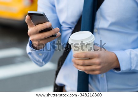 Close up of a businessman using mobile phone and holding paper cup. Close-up detail of a businessmanÃ¢??s hand holding paper cup and using a smartphone while walking on the road. Man going at work.