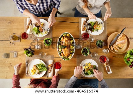 Top view of dining table with salad and roasted chicken with potatoes. High angle view of happy young friends having lunch at home. Men and women eating lunch together.