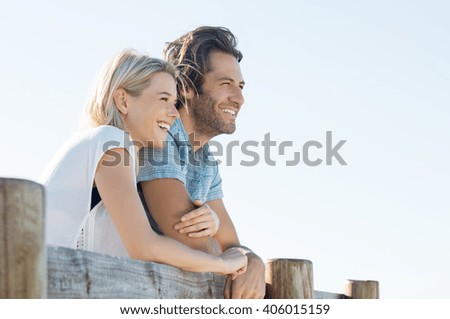 Happy young couple leaning over fence and looking away, copy space. Smiling couple thinking about the future. Young woman embracing her boyfriend outdoor during the sunshine.