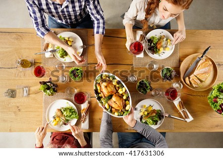 Top view of man passing food bowl to friend. High angle view of happy young friends eating together at home. Happy men and women having lunch with roasted chicken.