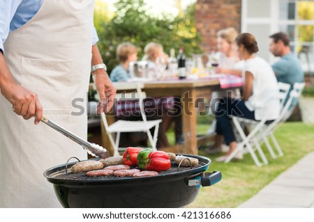Close up hands of a senior man barbequing in the garden with family in background. Grandfather cooking on grill. Man cooking hamburgers, sausages and peppers with barbeque.