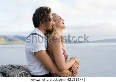 Couple hugging by the sea. Young couple at the beach holding each other. Boyfriend embracing girl from behind at beach during sunset at seashore.