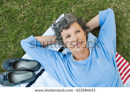 Pensive senior woman lying on towel on grass. High angle view of thoughtful mature woman thinking. Retired woman contemplating her life after retirement.