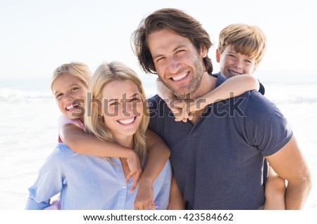 Parents giving piggyback ride to kids at beach. Close up of smiling family having fun at summer vacation. Portrait of happy family looking at camera at beach.