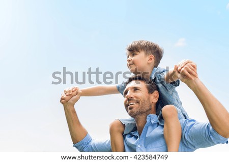 Father giving son ride on back in park. Portrait of happy father giving son piggyback ride on his shoulders and looking up. Cute boy with dad playing outdoor.