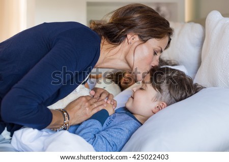 Mother giving good night kiss to sleeping son. Lovely mother putting son to bed. Happy young loving mother kisses son on forehead.