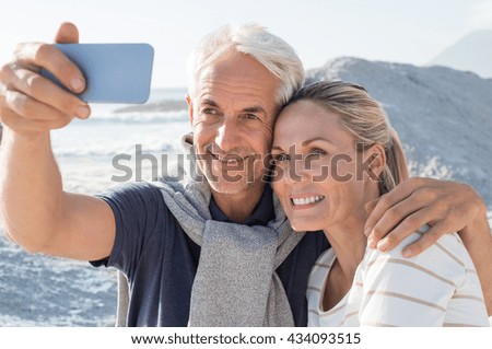 Happy romantic couple embracing on the beach and taking a photo with smart phone. Portrait of senior smiling couple taking a selfie in a summer vacation at sea. Happy mature couple take a picture.