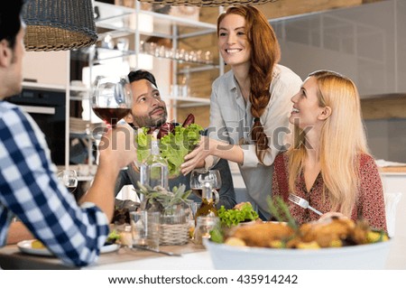 Host woman serving salad to guests at home. Smiling woman serving salads to her friends at home. Happy smiling people eating together for lunch. Group of friends enjoying meal at home together