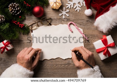 Top view of christmas letter in santa claus hand. Close up of hands holding empty wishlist on wooden table with xmas decoration. High angle view of santa claus hands writing on a paper with gifts.