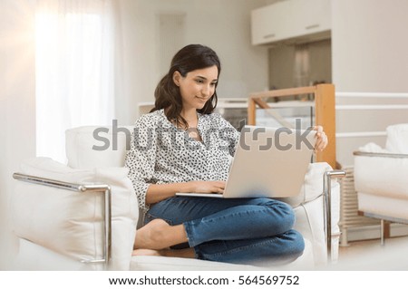 Young woman doing research work for her business. Smiling woman sitting on sofa relaxing while browsing online shopping website. Happy girl browsing through the internet during free time at home.