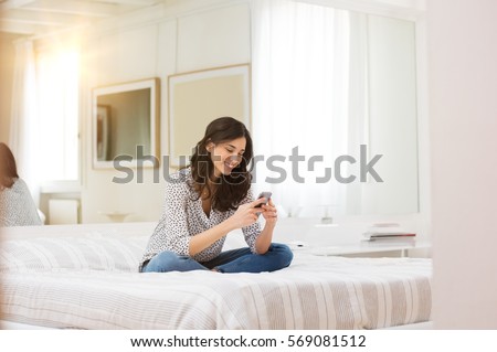Happy woman sitting on bed reading text message on smartphone. Smiling girl sitting in bedroom in casual in conversation with friend in a chat room. Smiling woman surfing net with cellphone at home.