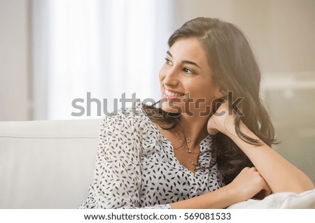 Confident woman relaxing on couch at home. Thoughtful smiling woman sitting on sofa and looking away. Portrait of satisfied girl daydreaming at home.