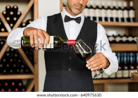 Close up of sommelier hands pouring red wine in a glass. Mature man in waistcoat pouring red wine in glass. Bartender serving wine in glass with shelfs of bottles behind.