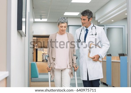 Mature doctor helping old female patient in crutches at the hospital. Physical therapist helping a woman on crutches in a medical clinic. Professional doctor and senior patient walking in hallway.