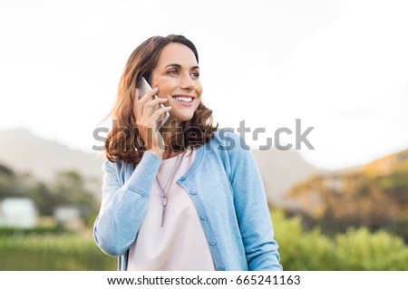 Mature happy woman talking on phone outdoor during sunset. Cheerful hispnic woman using smartphone and looking away. Happy woman in conversation using mobile phone and smiling.