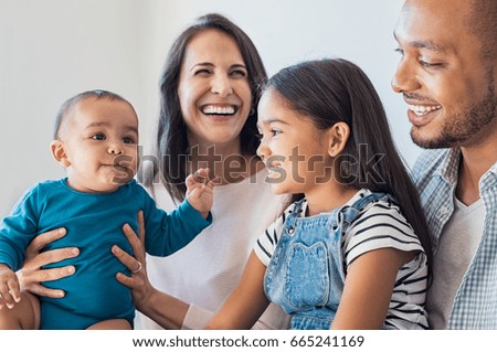 Happy daughter sitting on father legs and looking his little brother. Excited infant playing with elder sister. Portrait of smiling multiethnic family sit on sfoa and enjoying at home.