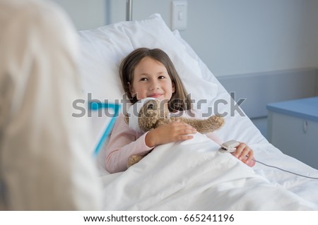 Smiling little girl with oxygen saturated probe resting on hospital bed. Girl patient looking at doctor with a smile. Child and doctor at medical clinic.