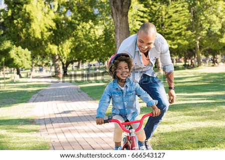 Lovely father teaching son riding bike at park. Happy father helping excited son to ride a bicycle at park. Young smiling black boy wearing bike helmet while learning to ride cycle with his dad.