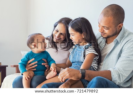 Multiethnic family playing with happy baby son at home. Parent and children relaxing together on the sofa at home in the living room. Little girl sitting on leg of dad looking her new cute brother.