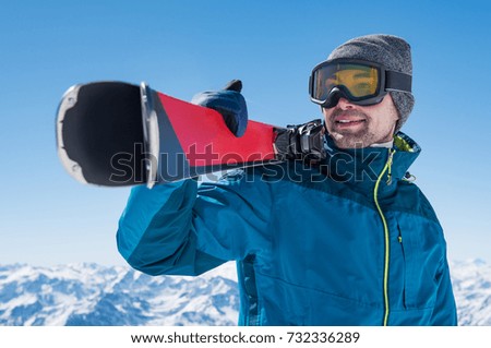 Happy skier holding a pair of skis and looking at the snowy mountains. Smiling guy with ski on shoulder standing in the mountain covered with snow. Man with winter equipment having fun.