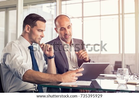 Businessman using a digital tablet to discuss information with senior leadership in a meeting. Business partners discussing plans. Mature boss and young business man working together in office.