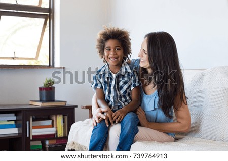 Happy latin mother embracing her adopted son on couch and smiling. Cheerful woman playing with her african child at home. Portrait of son and mom having fun together after school.