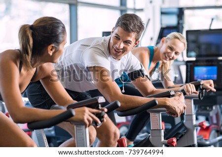 Young people talking and smiling while working out on bike at gym. Friends in a conversation while cycling on stationary bike in fitness centre. Group of happy people working out at spinning class.